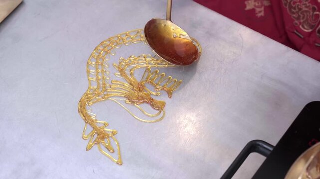 A near-complete tanghua sugar dragon glistens on a steel slab as the artisan adds final syrup threads with a bronze ladle, Thailand Chinese New Year fair. Part 4 of 6 &mdash; a tanghua sugar dragon created