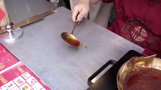 Close-up of a hand holding a bronze ladle, letting hot amber sugar syrup drip onto a steel slab before starting a tanghua sugar dragon, Thailand Chinese New Year festival. Part 1 of 6 tanghua sugar