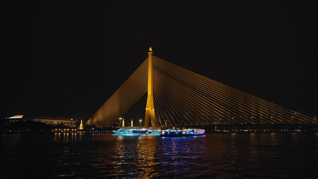 Illuminated tour boats cruise past as the cable-stayed span's golden lights power down, static wide shot, dramatic transitional night atmosphere