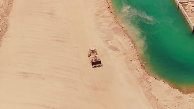 High angle view of a yellow bulldozer working on a construction site, moving and leveling sandy ground along the edge of a body of turquoise water, preparing the land for development