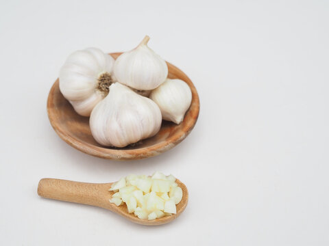 Chopped garlic is in a wooden spoon and garlic is in a wooden cup, placed on a white surface.