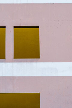 Across a flat wall, a white band divides the pink facade as yellow panels introduce geometric color, shaping minimal architecture into a measured pattern