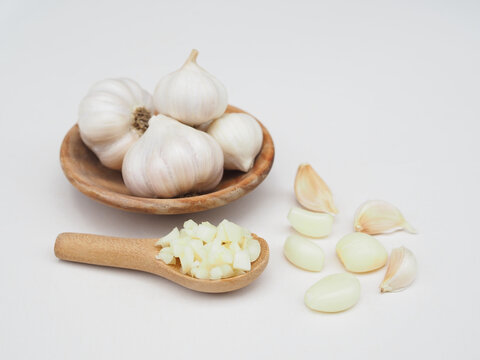Chopped garlic is in a wooden spoon and garlic is in a wooden cup, placed on a white surface.