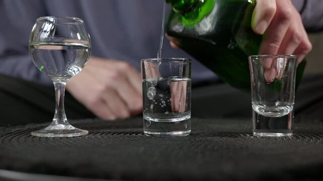 Bartender pouring strong alcoholic drink into shot glasses. Professional bartender pouring clear alcohol, like vodka or tequila, from a green bottle into two shot glasses on a black bar mat