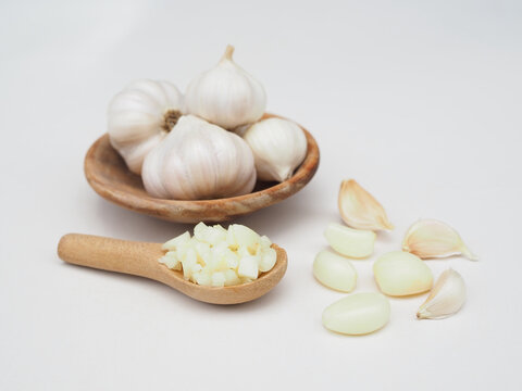 Chopped garlic is in a wooden spoon and garlic is in a wooden cup, placed on a white surface.