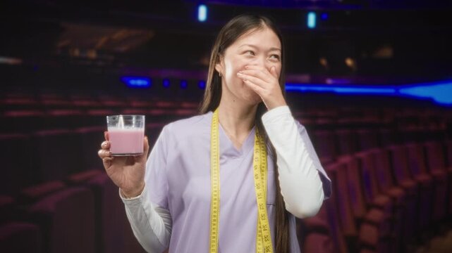 Woman doctor in lavender scrub holds pink beverage, hand covers mouth laughing, measuring tape around neck, auditorium; nutrition humor amusement.