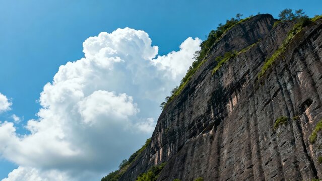 Steep rocky cliff face with green vegetation under a bright blue sky with large white clouds