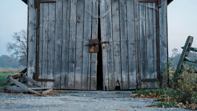 Rustic wooden barn doors with rusted iron hardware and a heavy latch, partially open, set in a rural landscape with fog during the early morning