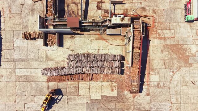 Workers are processing wood in an outdoor area. Large stacks of logs are lined up. Machinery is present, and signs of wood processing activity are evident on the ground.