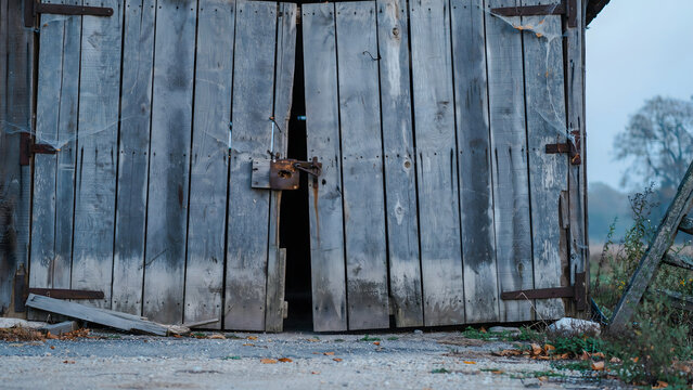 Weathered wooden barn doors with rusted metal hinges and a heavy iron latch, partially open, revealing a dark interior on a misty autumn morning