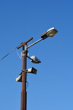 A low angle shot of a rusty metal utility pole with street lamps and public address horn speakers under a clear blue sky.