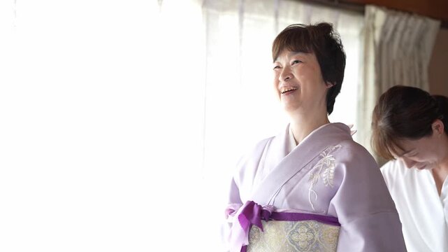 Soft light fills a quiet room. Woman adjusts a kimono's sash with care. Another smiles gently, observing the process. Tender moments captured in traditional attire. Perfect for wedding, cultural