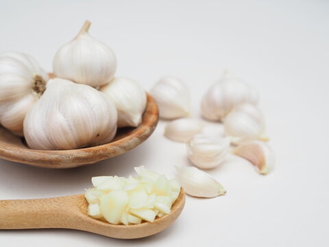 Chopped garlic is in a wooden spoon and garlic is in a wooden cup, placed on a white surface.