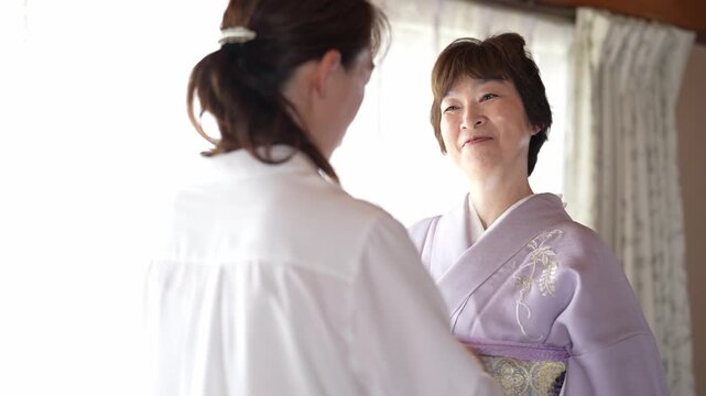 Soft light floods the room through sheer curtains. Two women share a quiet, tender moment. Younger woman gently adjusts older woman's kimono sash. Warmth radiates from their close
