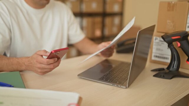 Man holding smartphone and paperwork with visible hand, checking laptop beside parcel boxes on a desk in building; focused entrepreneurship.