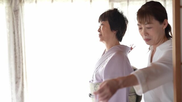 Soft light bathes a woman in traditional robes. She stands calmly, hands open, facing sideways. Another person leans forward nearby, partially out of frame. The quiet moment conveys peace