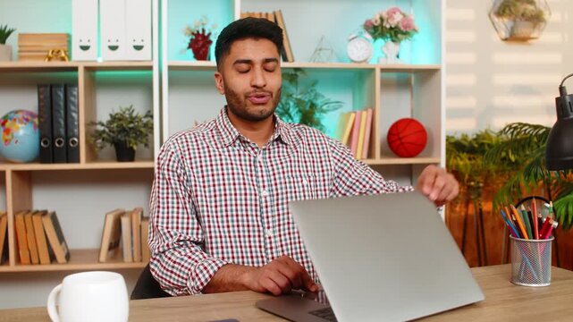 Indian man on home table closes laptop with satisfied smile after productive work and relief. Arabian guy on home table ends remote job with calm mood enjoying good results and daily progress indoors.