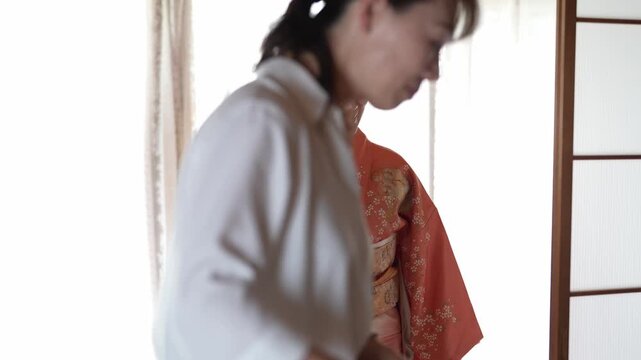 Soft light bathes a gentle scene. Woman adjusts a kimono with care. Another smiles warmly beside her. Traditional craftsmanship is tenderly cared for. Ideal for cultural, heritage, or bridal themes