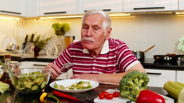 Senior old man tastes veggie salad in home kitchen feeling disappointed about dietary plan. Elderly grandfather pauses meal with fork thinking about weight loss nutrition plus healthy eating habits.