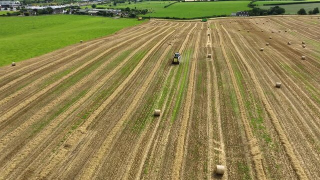 Tractor Pulling Round Baler Through Farmland With Village and Fields in Background