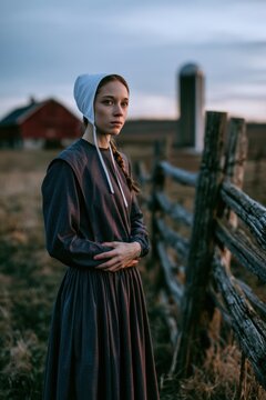 Amish woman in modest attire standing beside a barn and fields, rural portrait in calm daylight