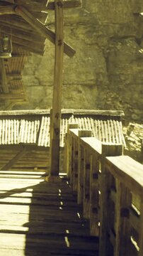 A weathered wooden bridge stretches across an ancient landscape, connecting to rustic buildings under the warm glow of sunlight. The canyon walls rise steeply around this serene spot.