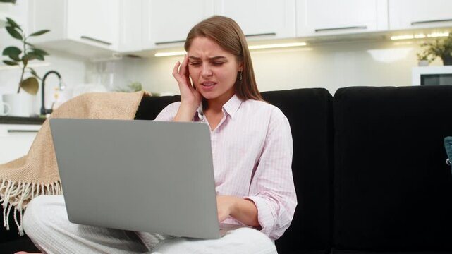 Young woman at home works on laptop holds temples headache squints and decides to rest to ease pain. Girl on sofa takes break closes eyes rubs head and neck and seeks calm after strong strain a bit.