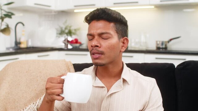 Indian man at home holds tea mug warms hands takes small sip relax mood and starts day with peace. Arabian guy on sofa enjoys hot drink focuses breathing and prepares mind for work with tea calm aim