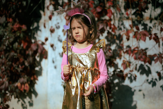 Annoyed Little Girl In Fairy Costume Squinting From Bright Sunshine. Disturbed child wearing butterfly wings blocks strong autumn sun glare with squinted eyes