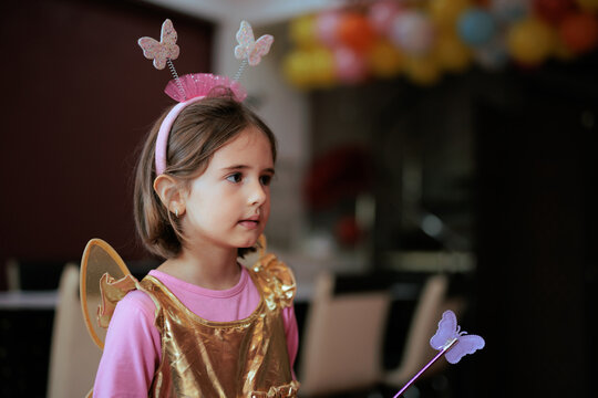 Happy Little Girl In Fairy Costume At Colorful Balloons Party. Cute child wearing golden dress and butterfly wings celebrates festive birthday event indoors