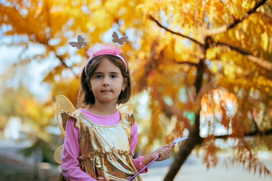 Little Girl In Butterfly Fairy Costume Holding Magic Wand Outdoors. Cute child wearing golden dress and wings celebrates autumn Halloween holiday in backyard