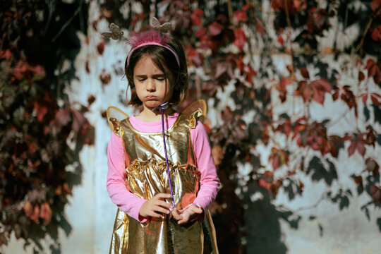 Annoyed Little Girl In Fairy Costume Squinting From Bright Sunshine. Disturbed child wearing butterfly wings blocks strong autumn sun glare with squinted eyes