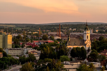 Panoramiczny widok na Ostrowiec Świętokrzyski i okolice w Polsce w letni wieczór. Na pierwszym planie widoczny jest kościół z zielonym dachem i wieżą. Wokół kościoła rosną bujne drzewa  © Grzegorz