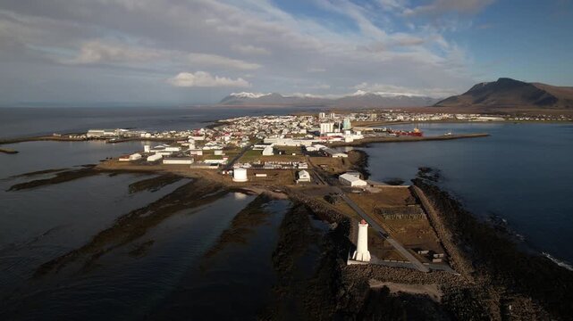 Stunning aerial footage of Akranes, Iceland, featuring a white lighthouse on rugged rocky shores, calm ocean waters, and snow-capped mountains under dramatic partly cloudy skies.