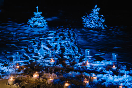 Atmospheric winter evening decor with glowing candles in glass cylinders placed on snowy ground, framed by illuminated fir trees. Ideal for romantic events or celebrations.