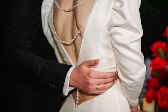 Romantic wedding moment, groom hugging bride from behind. Close-up view from the back, highlighting the bride's elegant white dress with a pearl necklace and the groom's wedding ring.