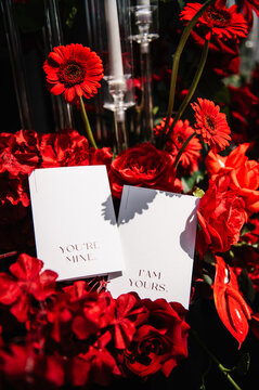 Romantic wedding place cards nestled among a vibrant display of red roses and gerbera daisies. The cards bear the sweet messages: "YOU'RE MINE" and "I AM YOURS.", adding a loving touch to event decor.