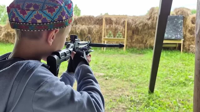 Boy wearing light blue hooded sweatshirt and bright skullcap is learning to shoot crossbow at cans with help of an adult instructor, an outdoor sports activity.