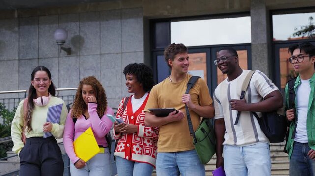 Happy multiracial group of college students laughing and talking while walking down the stairs of the campus building, showcasing friendship and the academic lifestyle in a university setting