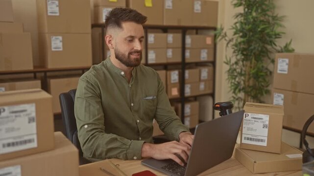 Man typing on laptop processing orders among shipping boxes with barcode scanner on desk in a building; productivity.