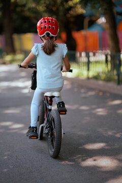 Little Girl Wearing Safety Helmet Riding Bicycle From Back View. Young child rides her small bike along concrete sidewalk outdoors practicing balancing skills.