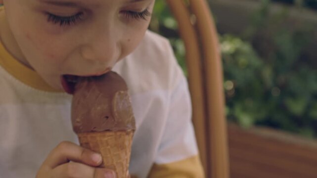 kid, boy, sitting on a woven chair outdoors and eating chocolate ice cream from a waffle cone. The child is holding the dessert with both hands during a sunny summer day.