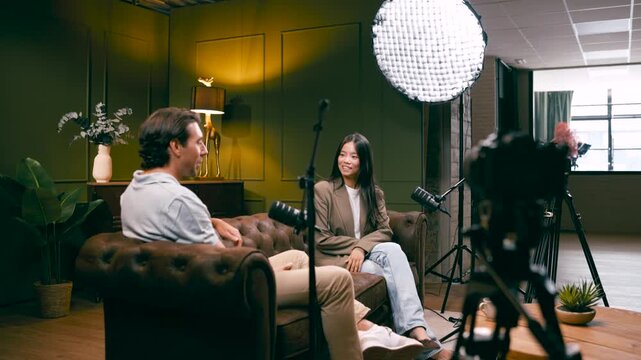 Young asian woman interviewing a caucasian man in a professional studio setup. Two podcasters are recording audio and video content for a show