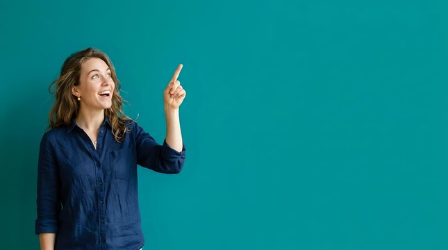 Young woman with long wavy brown hair and fair complexion wearing navy blue shirt pointing upward with excited expression against turquoise background for advertising and promotional use.