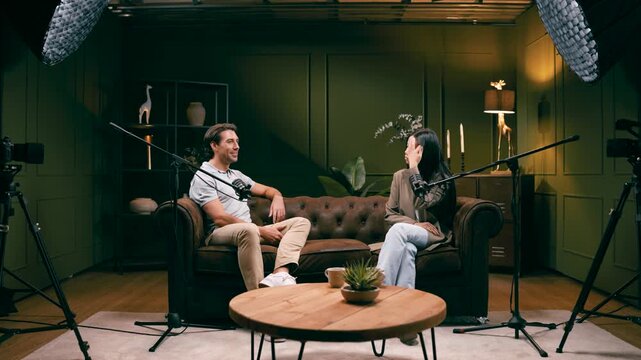 Two people, a man and a woman, having a conversation while sitting on a sofa. Professional studio setup with microphones and cameras