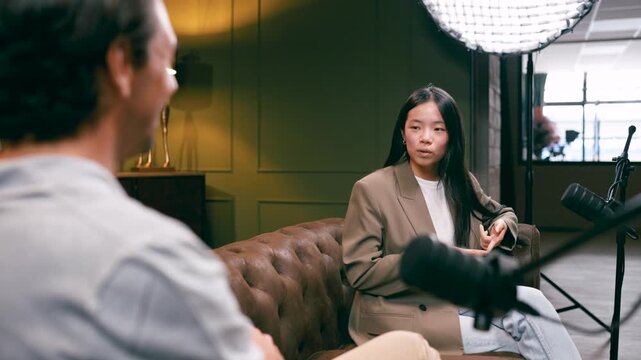 Over the shoulder view of a male host interviewing a young asian businesswoman. She actively gestures while talking into the microphone in a studio