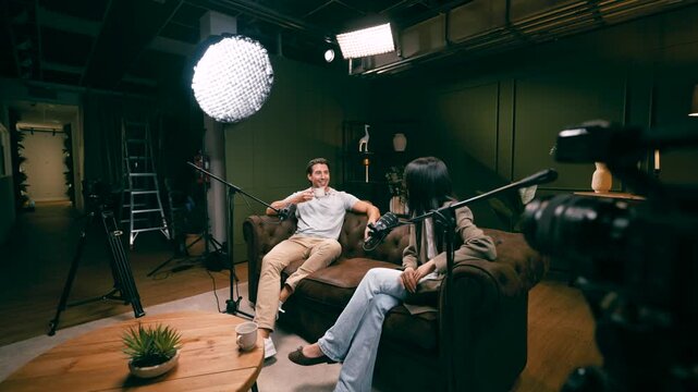 Male and female hosts recording a podcast in a professional studio. Two speakers sit on a sofa and talk into microphones during an interview