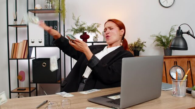 Young businesswoman at home office tosses dollar bills high laughing as money rain showers room. Freelancer girl at table imagines jackpot donates cash sharing wealth and celebrating generous fortune