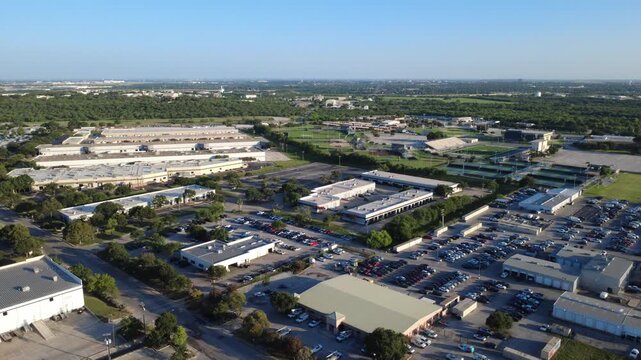 Industrial warehouses align near high school athletic complexes in Alamo Downs Industrial Park. Paved parking spaces border commercial buildings under clear morning skies, San Antonio