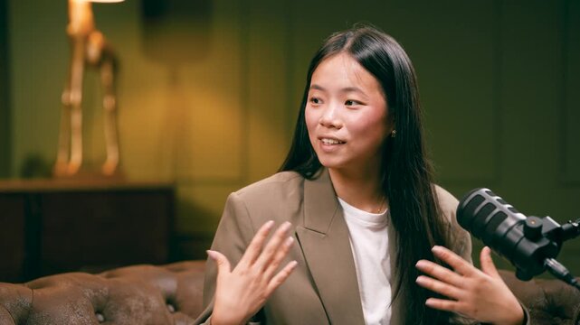 Charismatic young Asian woman smiling and gesturing while talking into a professional microphone during a podcast or live stream show